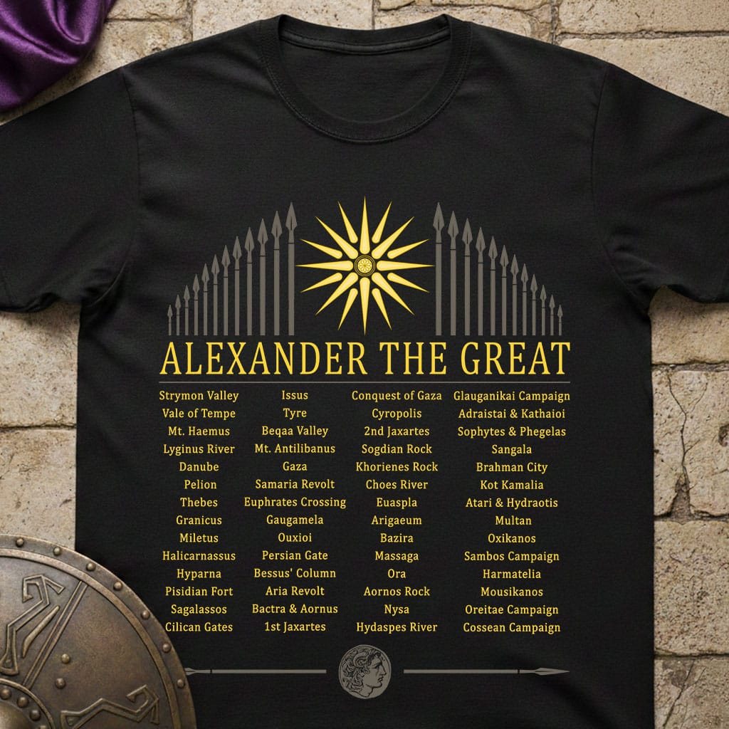 A black short-sleeve graphic T-shirt laid flat on a rustic stone surface, featuring a large gold sun and spearhead design with the text 'ALEXANDER THE GREAT' and a list of historical campaigns and battles in yellow and white lettering.