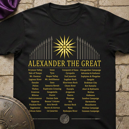 A black short-sleeve graphic T-shirt laid flat on a rustic stone surface, featuring a large gold sun and spearhead design with the text 'ALEXANDER THE GREAT' and a list of historical campaigns and battles in yellow and white lettering.