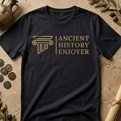 A black short-sleeve t-shirt with a gold-colored graphic that reads 'ANCIENT HISTORY ENJOYER' next to a classical column motif. The shirt is laid flat on a textured stone surface surrounded by decorative props like ancient coins, a ceramic jug, and dried leaves.