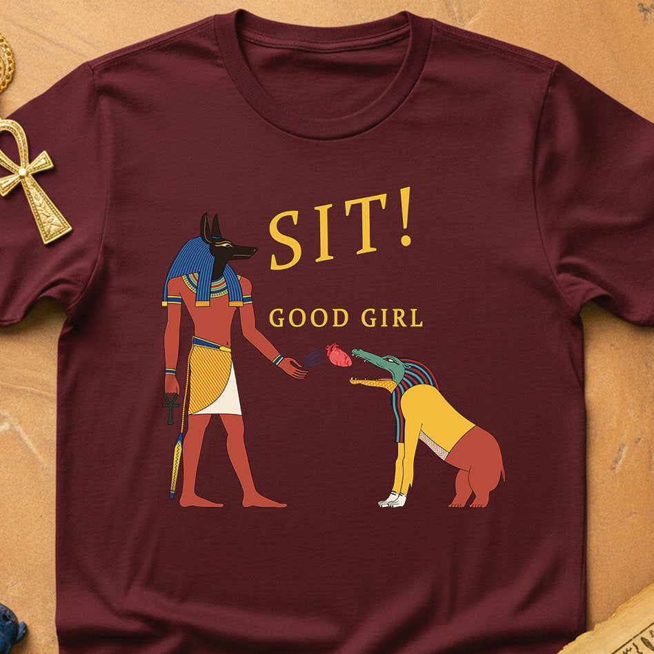 Maroon t-shirt with Egyptian-themed graphic and text 'SIT! GOOD GIRL' on a wooden surface.