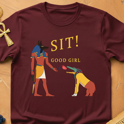 Maroon t-shirt with Egyptian-themed graphic and text 'SIT! GOOD GIRL' on a wooden surface.