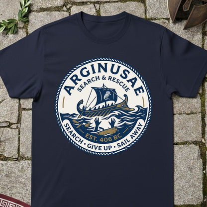 A navy blue crewneck t-shirt laid flat on a stone pavement background, featuring a large circular graphic print with a Viking ship, waves, and the text 'ARGINUSAE SEARCH & RESCUE'.