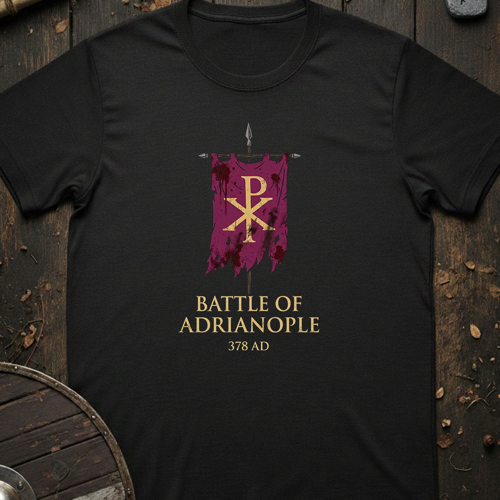 A black short-sleeve t-shirt laid flat on a rustic dark wood background, featuring a graphic of a purple banner with a gold Chi-Rho symbol and the text 'BATTLE OF ADRIANOPLE 378 AD' in gold.