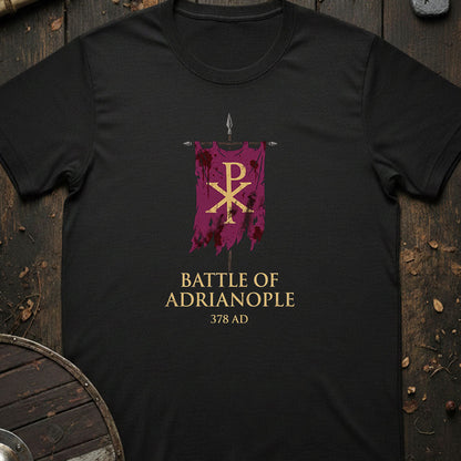 A black short-sleeve t-shirt laid flat on a rustic dark wood background, featuring a graphic of a purple banner with a gold Chi-Rho symbol and the text 'BATTLE OF ADRIANOPLE 378 AD' in gold.