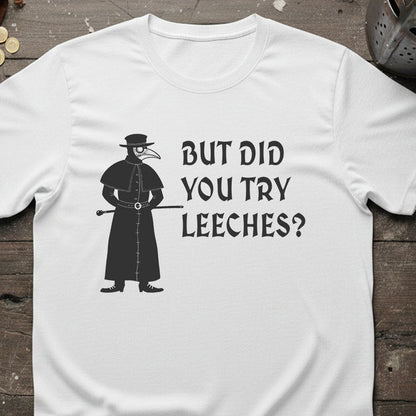 White t-shirt with a black silhouette of a plague doctor and text 'But did you try leeches?' on a wooden surface.