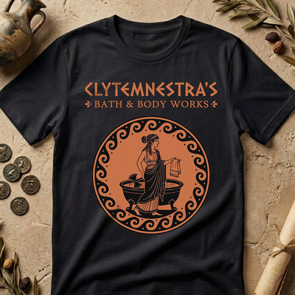 A black short-sleeve graphic T-shirt laid flat on a textured stone surface with decorative ancient props around it. The shirt has orange text reading 'CLYTEMNESTRA'S BATH & BODY WORKS' and a large circular orange-and-black illustration of a classical woman standing by a tub.