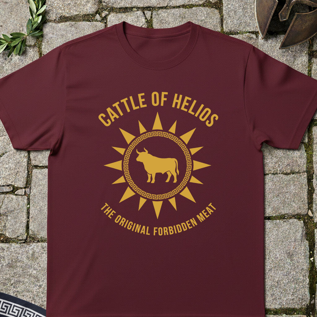 A maroon short-sleeve t-shirt with a gold graphic print reading 'CATTLE OF HELIOS' and 'THE ORIGINAL FORBIDDEN MEAT' around a bull silhouette, laid flat on a stone surface.