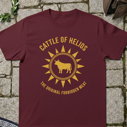 A maroon short-sleeve t-shirt with a gold graphic print reading 'CATTLE OF HELIOS' and 'THE ORIGINAL FORBIDDEN MEAT' around a bull silhouette, laid flat on a stone surface.
