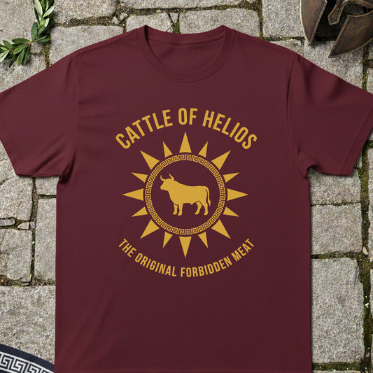 A maroon short-sleeve t-shirt with a gold graphic print reading 'CATTLE OF HELIOS' and 'THE ORIGINAL FORBIDDEN MEAT' around a bull silhouette, laid flat on a stone surface.