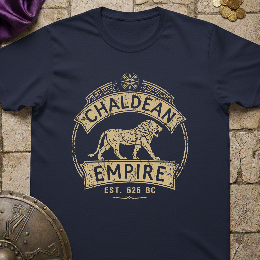 A navy blue short-sleeve t-shirt with a distressed beige graphic reading 'CHALDEAN EMPIRE EST. 626 BC' and featuring a lion illustration, laid flat on a stone-textured background with decorative props.