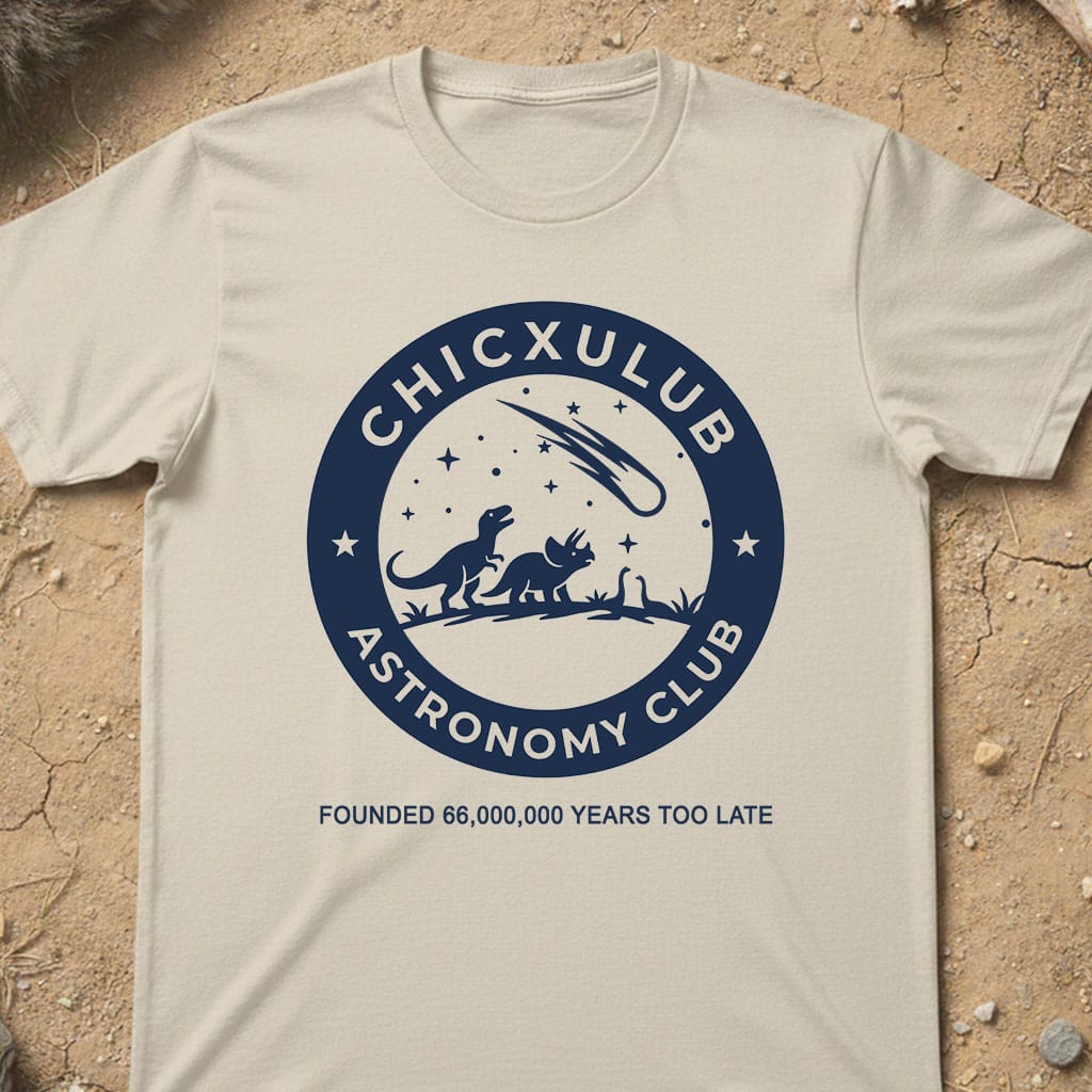A cream-colored short-sleeve t-shirt with a navy blue circular graphic reading 'CHICXULUB ASTRONOMY CLUB' and 'FOUNDED 66,000,000 YEARS TOO LATE', laid flat on a textured sandy surface.