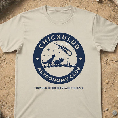 A cream-colored short-sleeve t-shirt with a navy blue circular graphic reading 'CHICXULUB ASTRONOMY CLUB' and 'FOUNDED 66,000,000 YEARS TOO LATE', laid flat on a textured sandy surface.