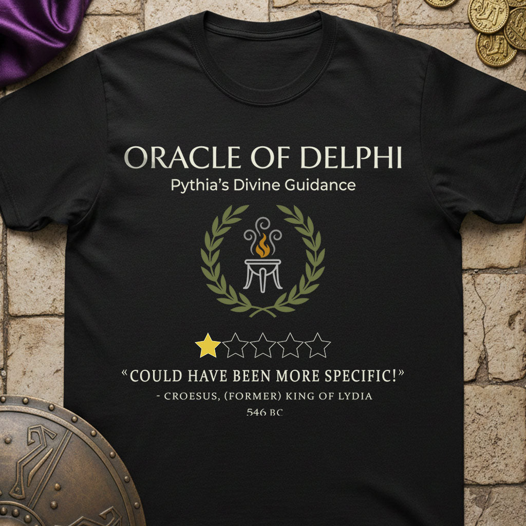 A black graphic t-shirt featuring a white and gold design with the text 'ORACLE OF DELPHI' and a laurel wreath around a tripod flame emblem, styled on a stone background with decorative props.