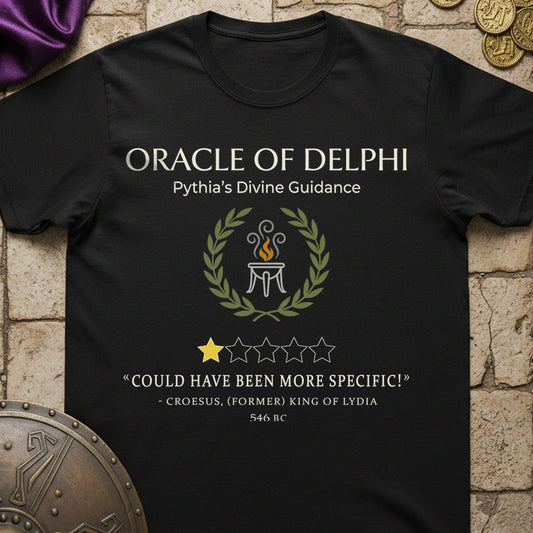 A black graphic t-shirt featuring a white and gold design with the text 'ORACLE OF DELPHI' and a laurel wreath around a tripod flame emblem, styled on a stone background with decorative props.