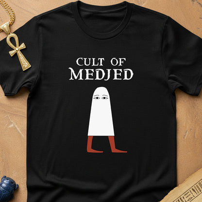 A black short-sleeve graphic t-shirt laid flat on a brown textured surface. The shirt features a white text print reading 'CULT OF MEDJED' above a stylized white figure with reddish-brown legs.