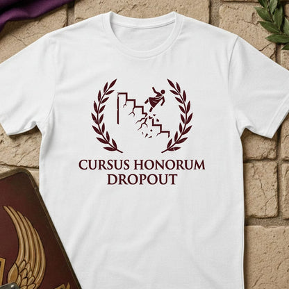 A white short-sleeve t-shirt with a dark red graphic print reading 'CURSUS HONORUM DROPOUT' under a laurel wreath and running figure design, displayed on a tan brick background with decorative props.