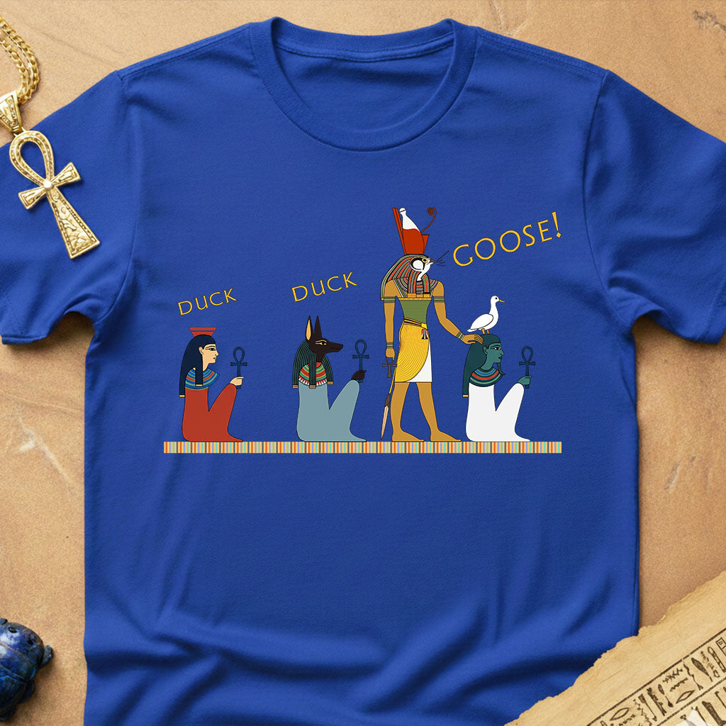 A royal blue short-sleeve t-shirt with a graphic print of Egyptian deities and the text 'DUCK', 'DUCK', and 'GOOSE!' in gold. The shirt is displayed flat on a textured tan background with decorative props including a gold ankh necklace and hieroglyphic paper.