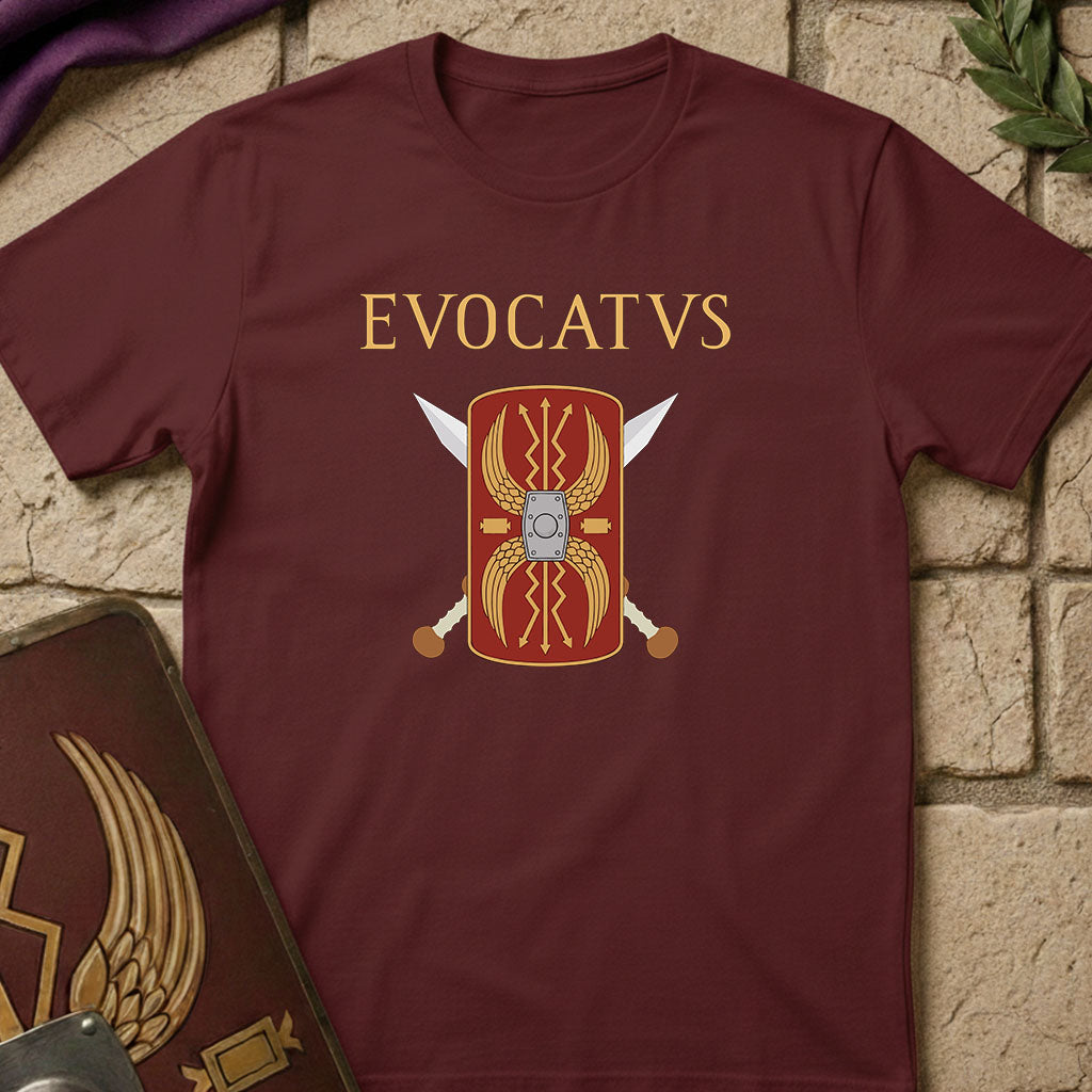 A maroon short-sleeve t-shirt with a graphic of a red and gold Roman shield, crossed swords, and the word 'EVOCATVS' in gold letters, displayed on a light brick background.