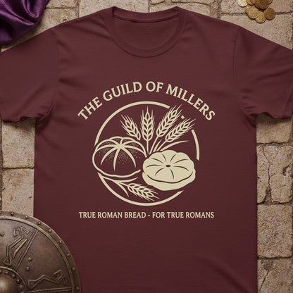 A maroon short-sleeve t-shirt with a cream-colored graphic reading 'THE GUILD OF MILLERS' and 'TRUE ROMAN BREAD - FOR TRUE ROMANS', featuring bread and wheat illustrations. The shirt is laid flat on a stone-textured background with Roman-themed props.