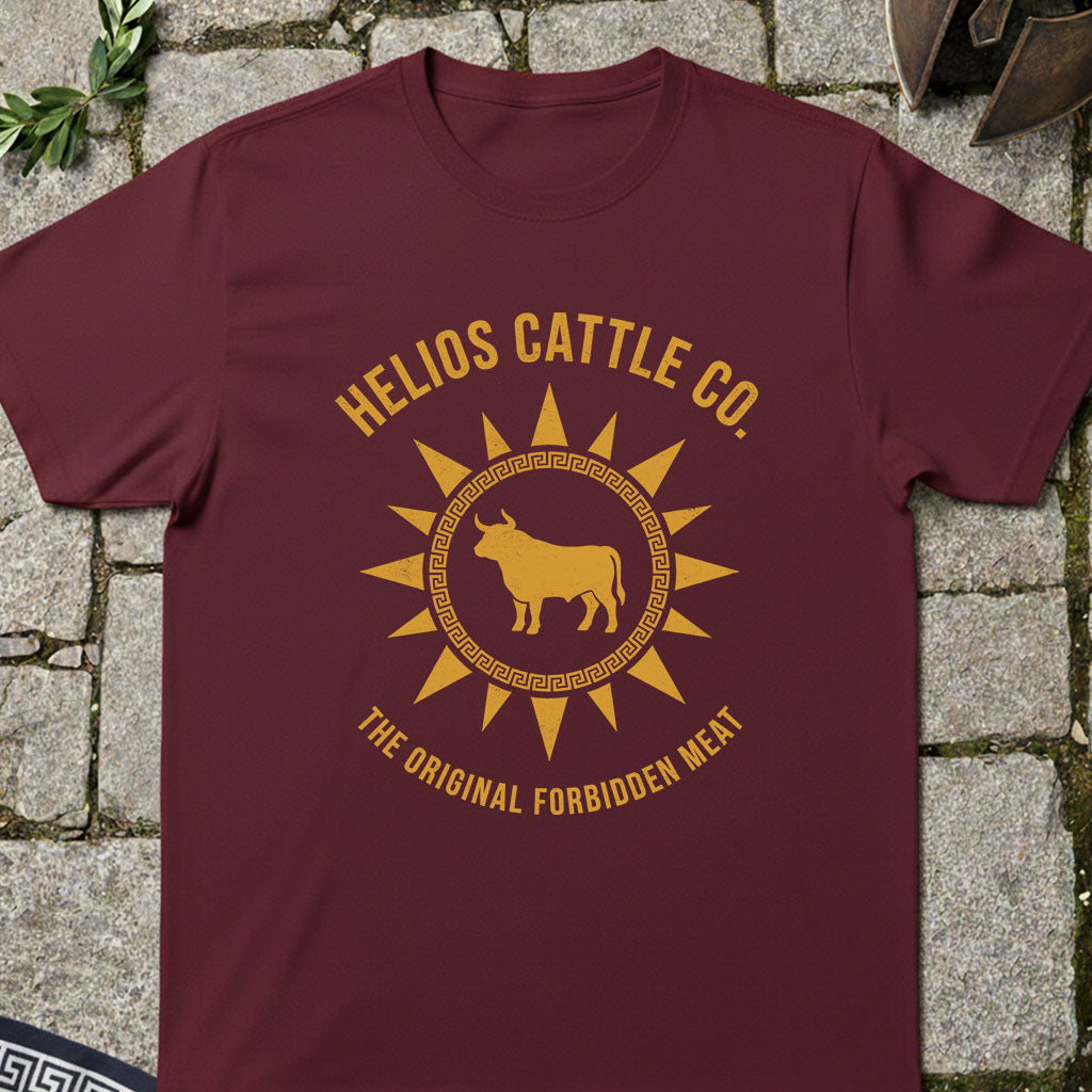 A maroon crewneck t-shirt with a golden-yellow graphic print reading 'HELIOS CATTLE CO.' and 'THE ORIGINAL FORBIDDEN MEAT' around a bull inside a sun design, laid flat on a gray stone background.