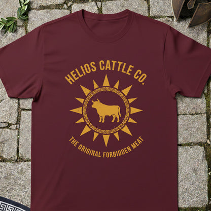 A maroon crewneck t-shirt with a golden-yellow graphic print reading 'HELIOS CATTLE CO.' and 'THE ORIGINAL FORBIDDEN MEAT' around a bull inside a sun design, laid flat on a gray stone background.