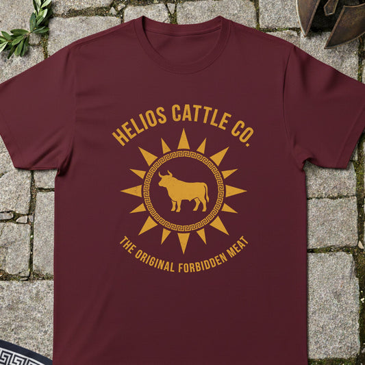 A maroon crewneck t-shirt with a golden-yellow graphic print reading 'HELIOS CATTLE CO.' and 'THE ORIGINAL FORBIDDEN MEAT' around a bull inside a sun design, laid flat on a gray stone background.