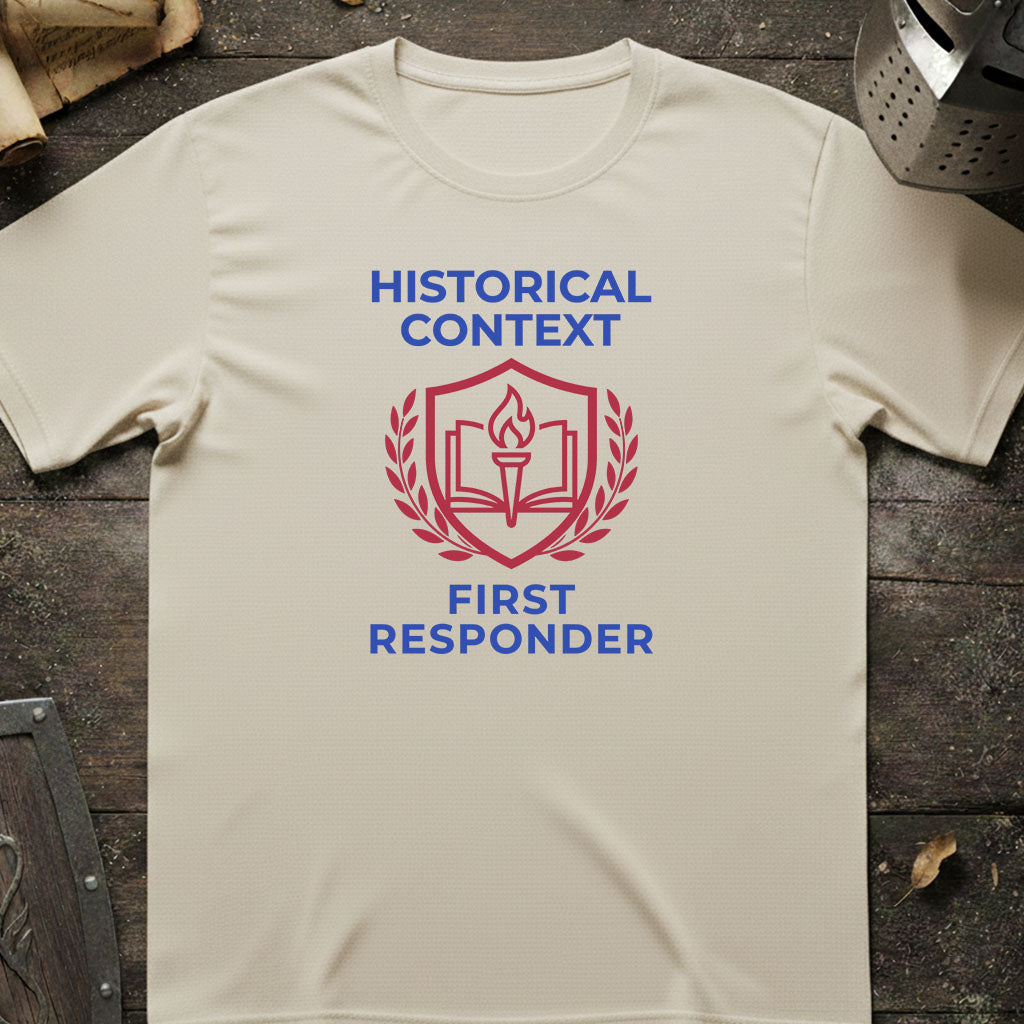 A cream-colored short-sleeve t-shirt with a red and blue graphic on the chest reading 'HISTORICAL CONTEXT FIRST RESPONDER' around an open book and shield emblem, laid flat on a dark rustic wooden surface.