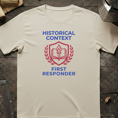 A cream-colored short-sleeve t-shirt with a red and blue graphic on the chest reading 'HISTORICAL CONTEXT FIRST RESPONDER' around an open book and shield emblem, laid flat on a dark rustic wooden surface.