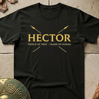 A black short-sleeve t-shirt with a gold graphic print reading 'HECTOR PRINCE OF TROY - Tamer of Horses' with crossed arrows, laid flat on a textured stone surface with decorative bronze props.