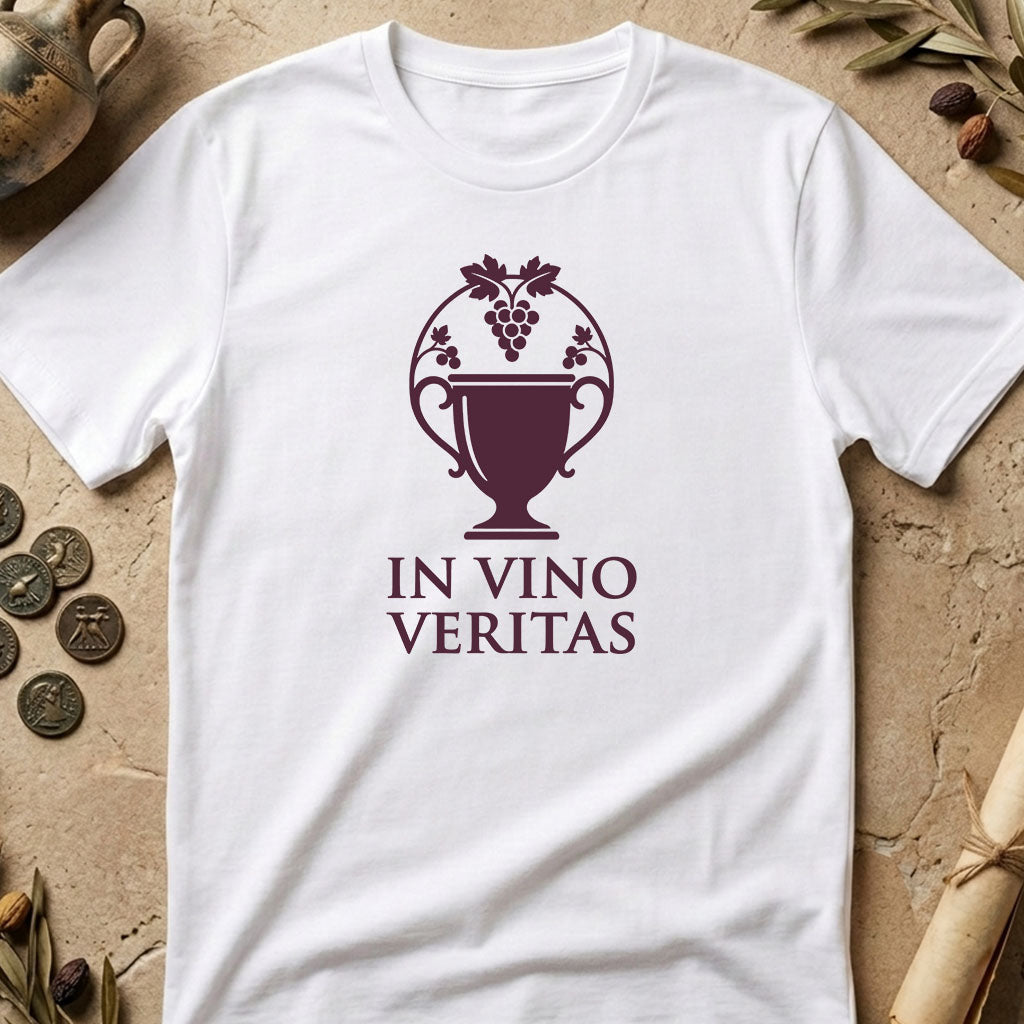A white short-sleeve T-shirt laid flat on a textured beige surface with decorative props around it. The shirt has a dark burgundy graphic of a wine cup with grapes and the words 'IN VINO VERITAS' printed on the front.