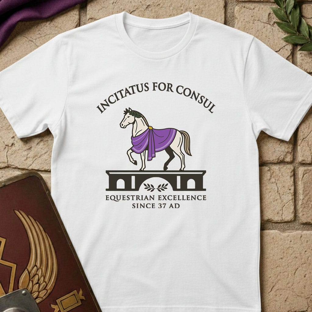 A white short-sleeve graphic T-shirt laid flat on a tan brick surface, with a printed design showing a horse draped in purple and black text reading 'INCITATUS FOR CONSUL' and 'EQUESTRIAN EXCELLENCE SINCE 37 AD'. Decorative props are partially visible at the edges.