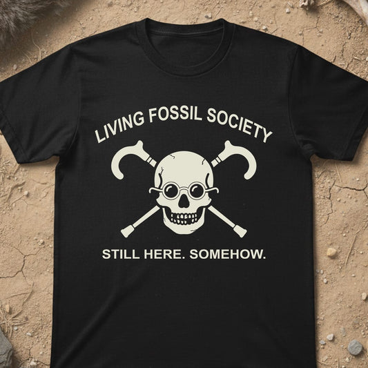 A black short-sleeve t-shirt with a white graphic print of a skull wearing glasses and crossed tools, with the text 'LIVING FOSSIL SOCIETY' and 'STILL HERE. SOMEHOW.