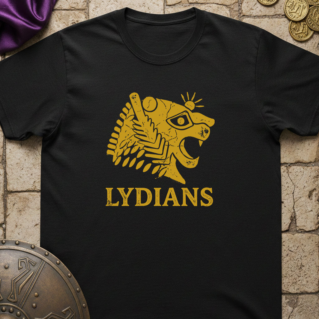 A black short-sleeve t-shirt with a gold graphic of a stylized lion head and the word 'LYDIANS' printed on the front, displayed flat on a stone-textured background with decorative props.