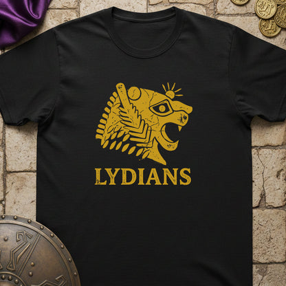 A black short-sleeve t-shirt with a gold graphic of a stylized lion head and the word 'LYDIANS' printed on the front, displayed flat on a stone-textured background with decorative props.