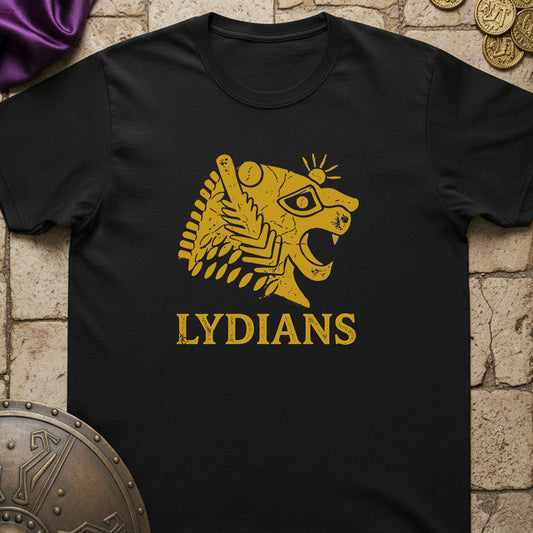A black short-sleeve t-shirt with a gold graphic of a stylized lion head and the word 'LYDIANS' printed on the front, displayed flat on a stone-textured background with decorative props.