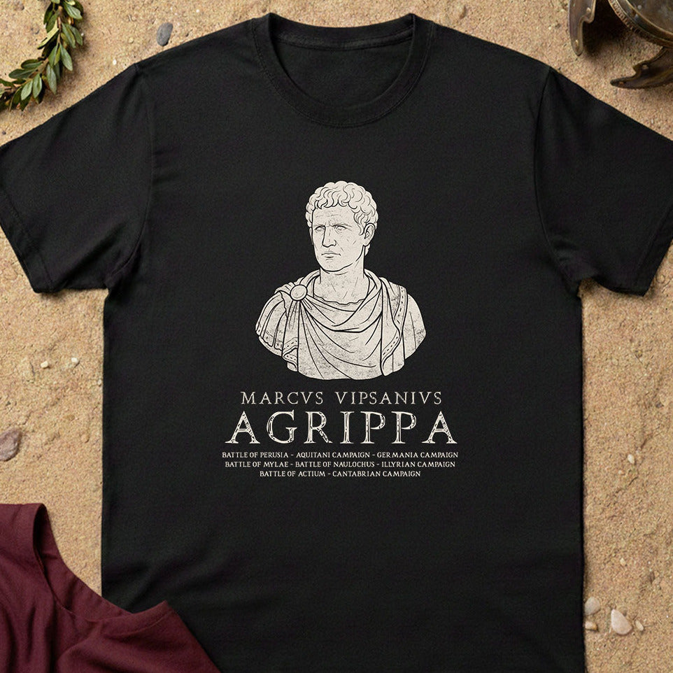 A black short-sleeve graphic T-shirt laid flat on a sandy surface with small decorative props around it. The shirt features a white line drawing of a classical bust above the text 'MARCUS VIPSANIUS AGRIPPA' and smaller battle-related text.
