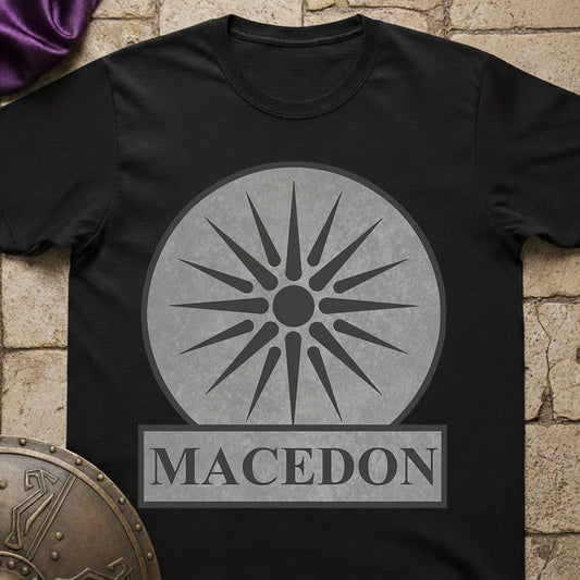 Black t-shirt with a gray sun-like symbol and 'Macedon' text on a stone surface.