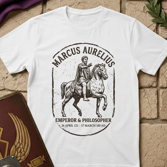 A white short-sleeve graphic T-shirt laid flat on a tan stone-textured background, with a vintage-style black print reading “MARCUS AURELIUS” and “EMPEROR & PHILOSOPHER” around an illustration of a rider on a horse.