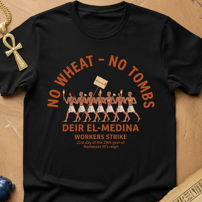 A black graphic t-shirt laid flat on a textured tan background with decorative props including an Egyptian ankh necklace, a blue stone, and paper. The shirt features an orange and brown graphic of workers marching with a sign, accompanied by text reading 'NO WHEAT - NO TOMBS' and 'DEIR EL-MEDINA WORKERS STRIKE'.