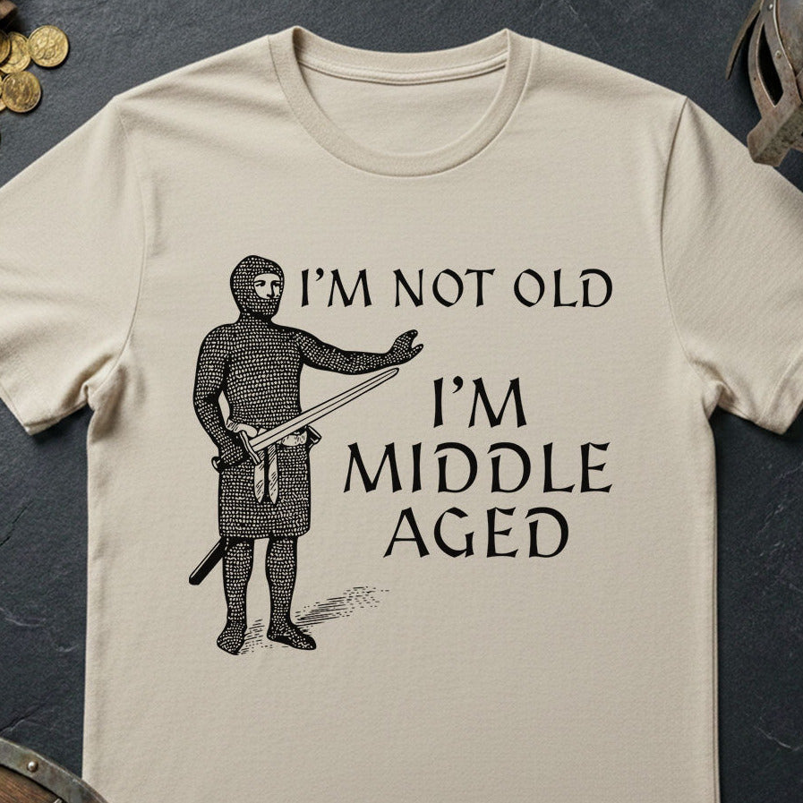 Beige t-shirt with a graphic of a person in medieval armor and text 'I'm not old, I'm middle aged' on a dark surface.