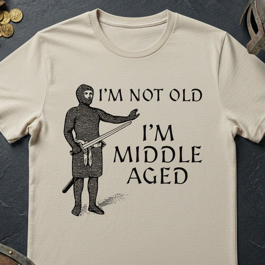 Beige t-shirt with a graphic of a person in medieval armor and text 'I'm not old, I'm middle aged' on a dark surface.