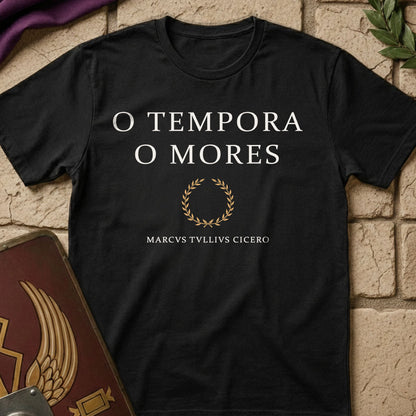 A black short-sleeve t-shirt with white text reading 'O TEMPORA O MORES' and a gold laurel wreath graphic, laid flat on a beige brick surface with decorative props.
