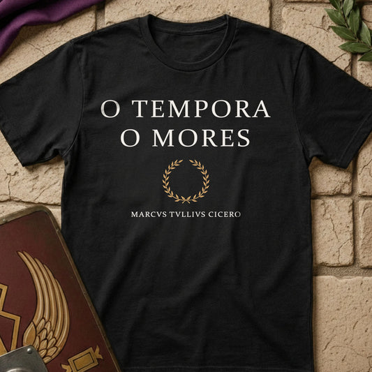 A black short-sleeve t-shirt with white text reading 'O TEMPORA O MORES' and a gold laurel wreath graphic, laid flat on a beige brick surface with decorative props.