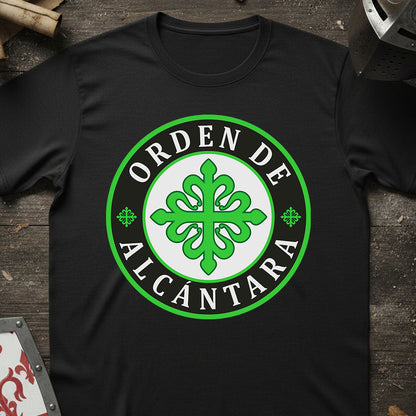 A black short-sleeve t-shirt laid flat on a dark rustic wooden surface, featuring a large circular green and white graphic on the chest that reads 'ORDEN DE ALCANTARA' around a green ornamental emblem.