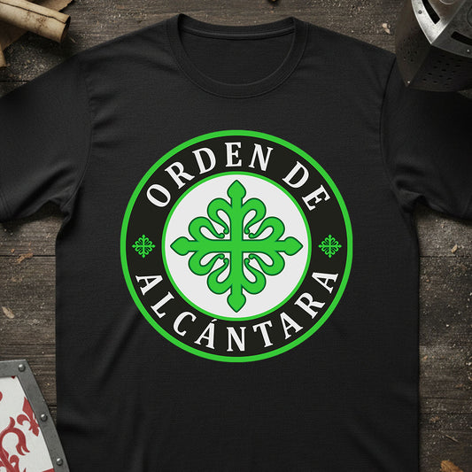A black short-sleeve t-shirt laid flat on a dark rustic wooden surface, featuring a large circular green and white graphic on the chest that reads 'ORDEN DE ALCANTARA' around a green ornamental emblem.