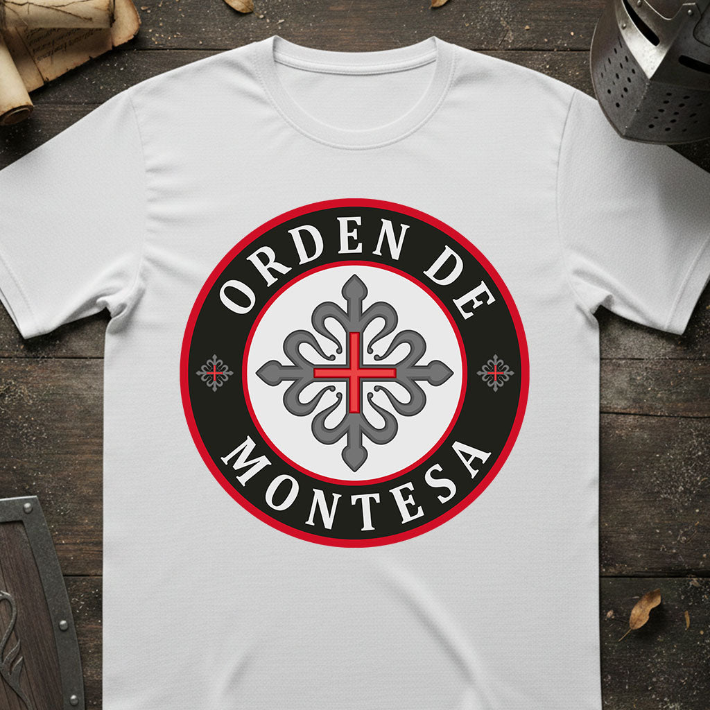 A white short-sleeve t-shirt laid flat on a dark rustic wooden surface, featuring a circular graphic print with the text 'ORDEN DE MONTESA' in white lettering on a black ring with a red border, surrounding a gray ornamental cross emblem.