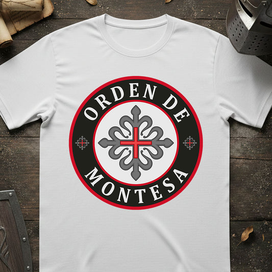 A white short-sleeve t-shirt laid flat on a dark rustic wooden surface, featuring a circular graphic print with the text 'ORDEN DE MONTESA' in white lettering on a black ring with a red border, surrounding a gray ornamental cross emblem.
