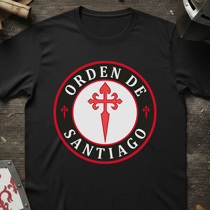 A black short-sleeve t-shirt laid flat on a rustic dark wood surface, featuring a large circular graphic on the chest with the words 'ORDEN DE SANTIAGO' in white around a red and white central emblem.