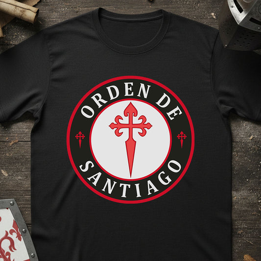 A black short-sleeve t-shirt laid flat on a rustic dark wood surface, featuring a large circular graphic on the chest with the words 'ORDEN DE SANTIAGO' in white around a red and white central emblem.