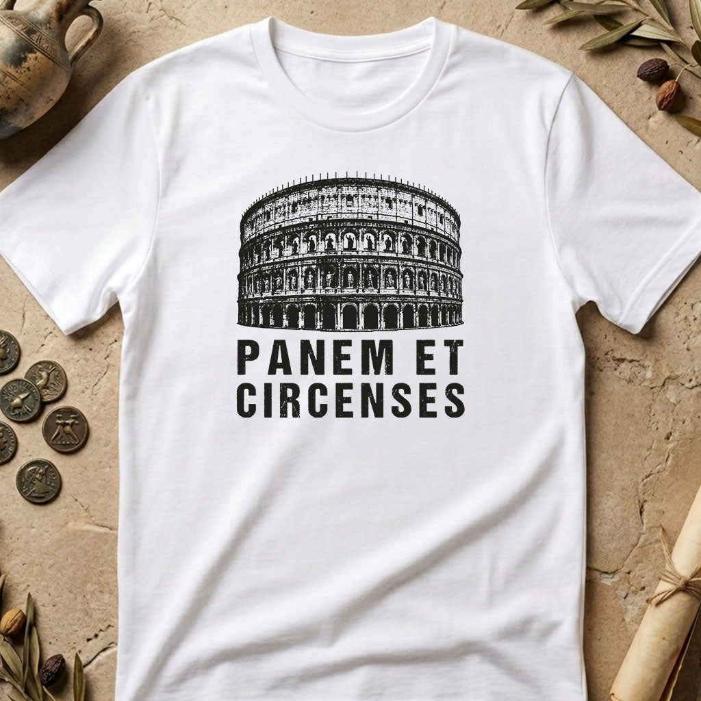 A white short-sleeve T-shirt laid flat on a textured beige surface with decorative props including coins, dried leaves, and rolled parchment. The shirt has a black graphic of the Colosseum and the text 'PANEM ET CIRCENSES' printed on the front.